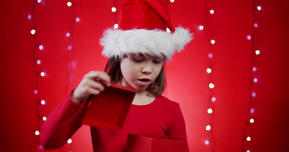 Cute Girl in a Santa Hat Opens a Gift on a Red Christmas Background