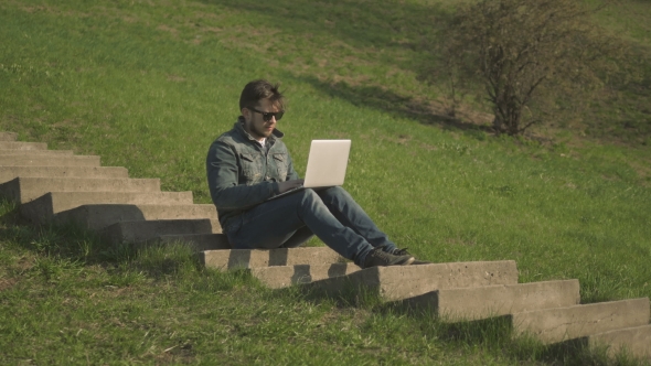 Young Hipster Man In Sunglasses Sitting On The Stairs Using Laptop