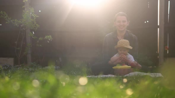 Happy Young Cheerful Mother Holding Baby Eating Fruits On Green Grass