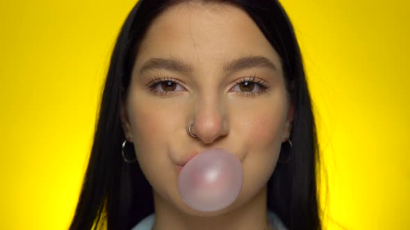 Attractive Woman Looking at Camera and Blowing Bubble Gum on Yellow Background
