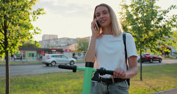 Happy woman stands with modern electric scooter talking on phone in city