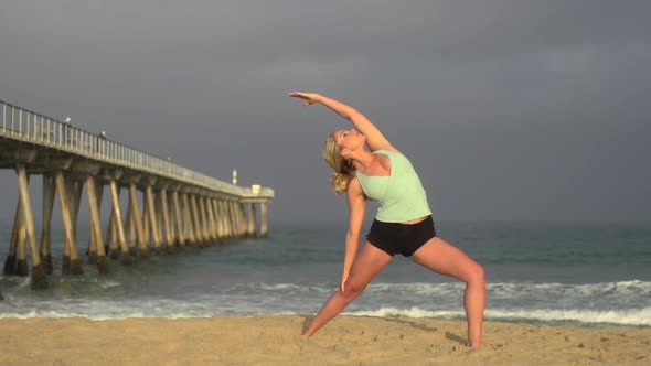 A young woman does yoga on the beach next to a pier.