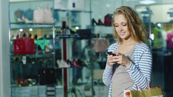 Attractive Female Shopper With Shopping Bags Uses a Smartphone