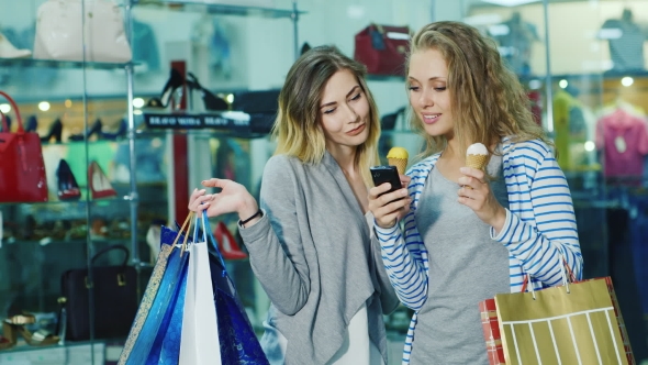 Two Young Women With Shopping Bags Dyal Go Ice-cream And Consider Something On The Phone. Smile, It