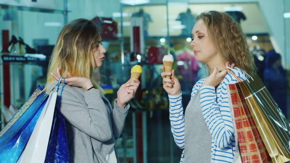 Two Friends Eating Ice Cream. Standing With Shopping Bags In a Store In The Background Of Glass