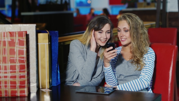 Relax After a Successful Shopping Experience. Two Women Sitting At a Table And View Something On