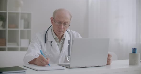 Elderly Doctor Is Listening Patient at Online Consultation, Looking at Screen of Laptop and Writing