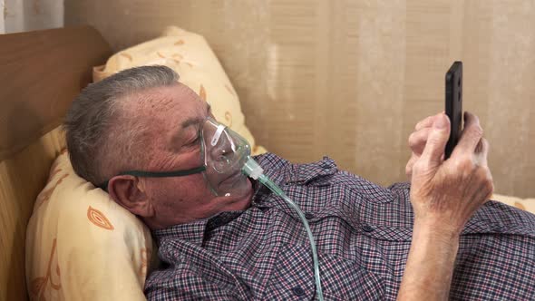 A Senior Man in Oxygen Mask Is Lying on Bed and Watching Something in His Smartphone