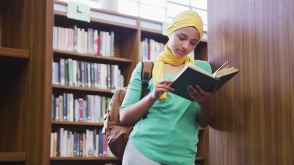 An Asian female student wearing a yellow hijab leaning against bookshelves and reading a book