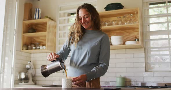 Happy caucasian woman standing in cottage kitchen pouring coffee from pot and smiling