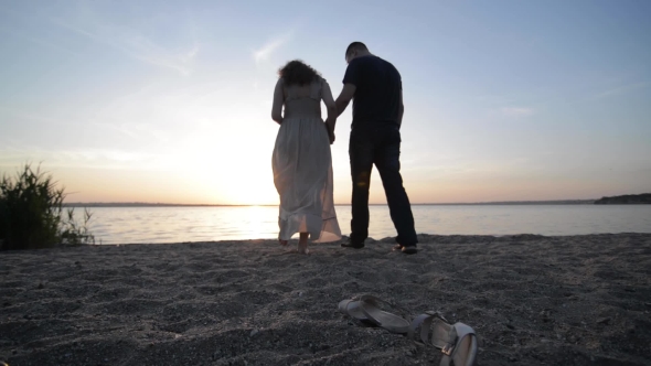 Couple Expexting Baby Walking On a River Shore Holding Hands On Sunset