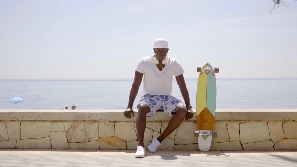 Skater Seated On Stone Wall By Ocean On Sunny Day