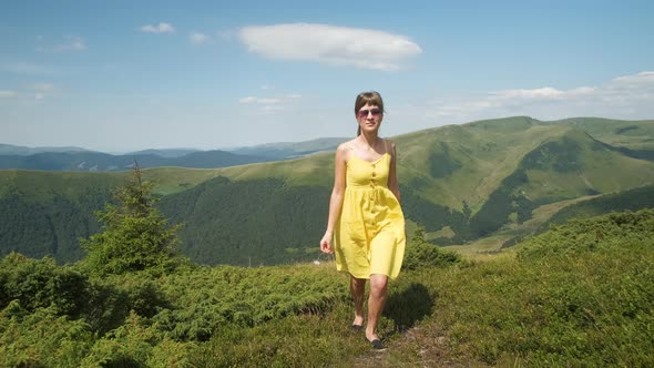 Young Happy Woman in Yellow Dress Walking on Grass Field on a Windy Day in Summer Mountains