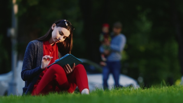 Stylish Woman Sitting On The Grass In The Park, Enjoying The Tablet alt
