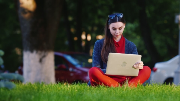 Female Student Sitting On The Grass In The Park, Uses a Laptop