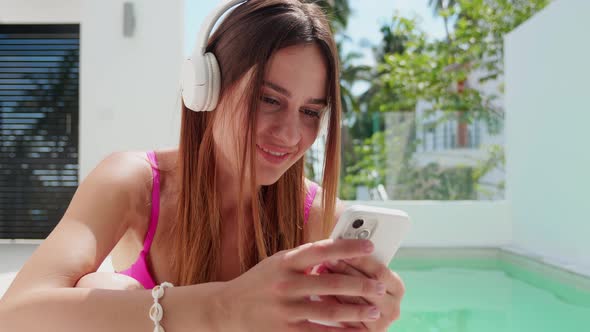Closeup of a Girl in Headphone in a Swimsuit Hold Phone on the Background of the Pool