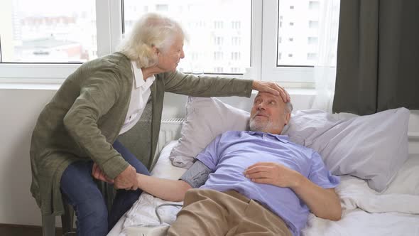 Caring Elderly Woman Checks the Blood Pressure of Her Old Man with Hypertension While Lying at Home