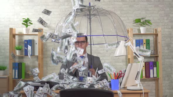 Indifferent Businessman with an Umbrella Sits at Desk in the Office and Slow Banknotes Rain Down on