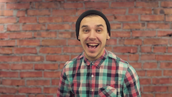 Blogger In Black Hat, Plaid Shirt Pronounce Words In Camera. Smiling. Brick Wall On Background.