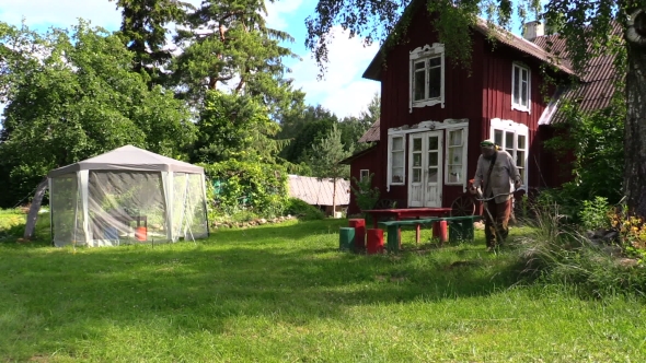 Gardener Man Cut Grass With Trimmer Near Rural House And Bower