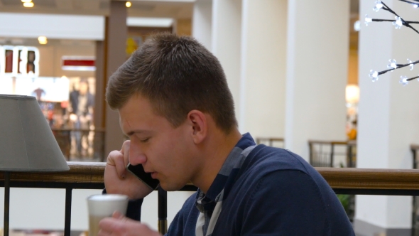 Young Handsome Man Drinking Coffee And Talking On The Cellphone In The Cafe In Shopping Mall.