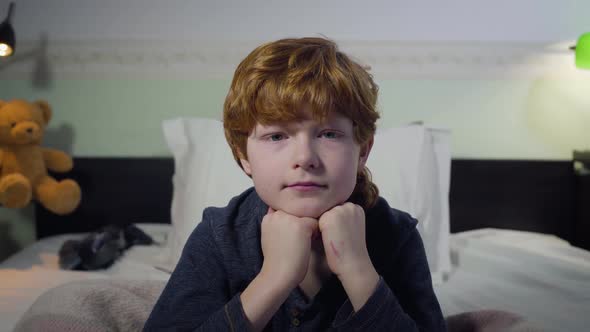 Headshot of Cute Caucasian Redhead Kid Sitting on Bed and Looking at Camera. Portrait of Little Boy