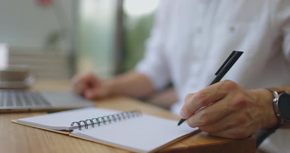 Man's Hands Writing Notes in Notebook with Black Ball Pen Sitting with Laptop at Wooden Cafe Table