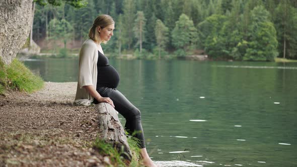 Pregnant woman touching gently her tummy when sitting near the mountain lake