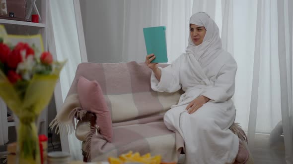 Happy Smiling Middle Eastern Woman in White Hijab and Bathrobe Waving at Tablet Talking Sitting on