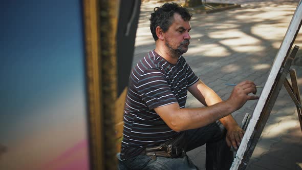 Male Artist Drawing Picture on Canvas Using Oil Paints While Sitting on an Alley of City Park