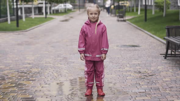 Portrait of Cute Little Girl in Pink Waterproof Costume and Rubber Boots Standing in Puddle and