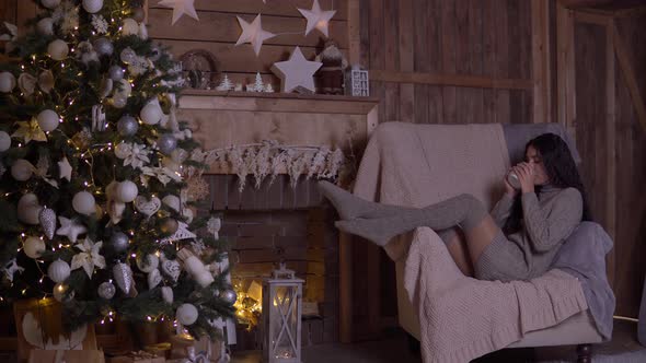 A Girl Drinks Tea Sitting in a Chair Near the Christmas Tree on Christmas Day