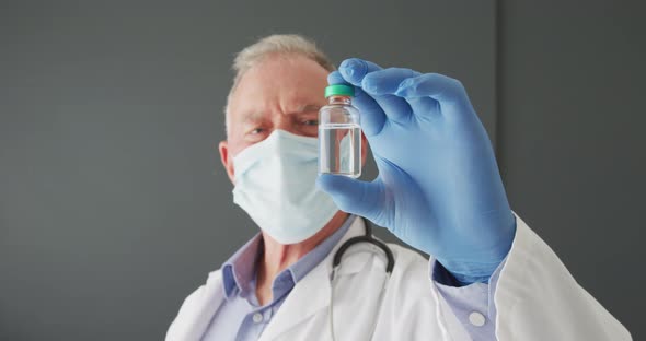 Portrait of caucasian senior male doctor wearing face mask holding covid-19 vaccine