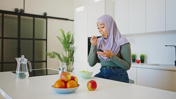 Busy Modern Arab Woman Eating Salad in the Kitchen and Looking at Her Smartphone Screen