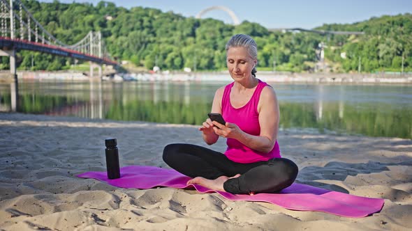 Senior Woman Sits on Pink Mat Looking in Smartphone By River