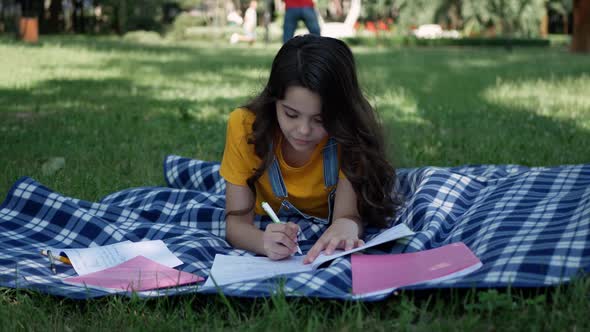 Little girl lying on a blanket. Cheerful girl lying down teaches lessons outdoors