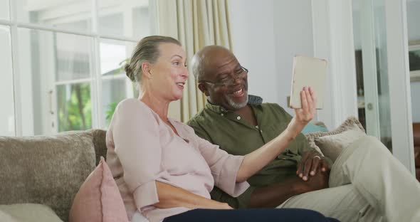 Happy senior diverse couple wearing shirts and using tablet in living room