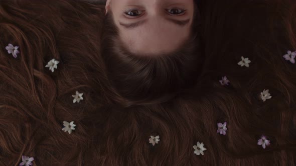 Woman with Flowers in Her Loose Hair Lies and Looks at the Camera Blinking