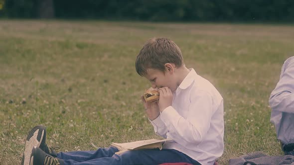 Young Concentrated Pupil Reads Book Eating Burger on Lawn