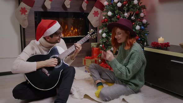 Man wearing santa hat playing the guitar with woman next to the fireplace.