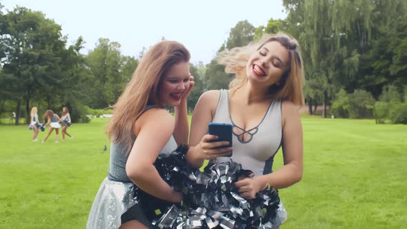 Two Cheerleading Girls Having Fun Surfing Internet on Smartphone and Laughing Standing Outdoors