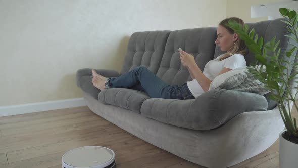 Attractive Girl Using Smartphone While Robotic Vacuum Cleaner Cleaning House