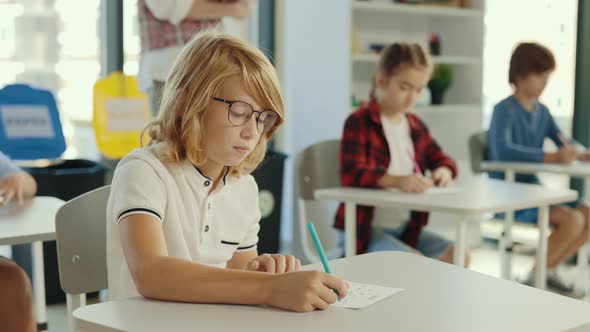 Redhead Pupil Wearing Glasses Sits at a Desk at School and Fills Out Tests and the Teacher in the