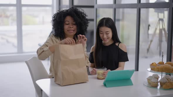 Young Smiling African American and Asian Women Unpacking Takeaway Food Sitting in Home Office at