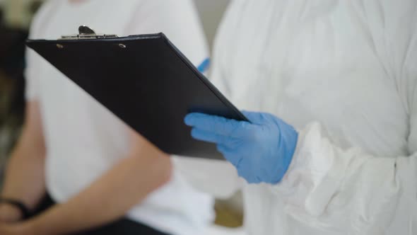 Unrecognizable Doctor in Antiviral Suit and Protective Gloves Taking Medical History of Patient in