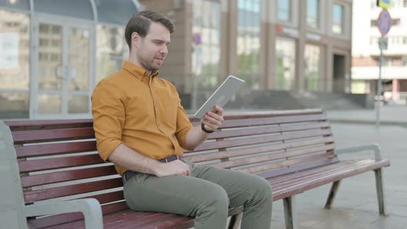 Online Video Chat on Tablet by Young Man Sitting Outdoor on Bench