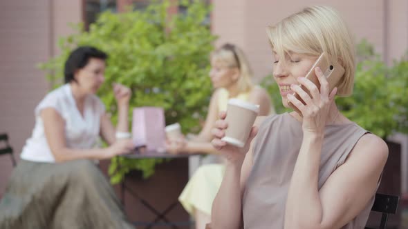 Portrait of Relaxed Joyful Blond Woman Talking on the Phone and Drinking Coffee in Outdoor Cafe