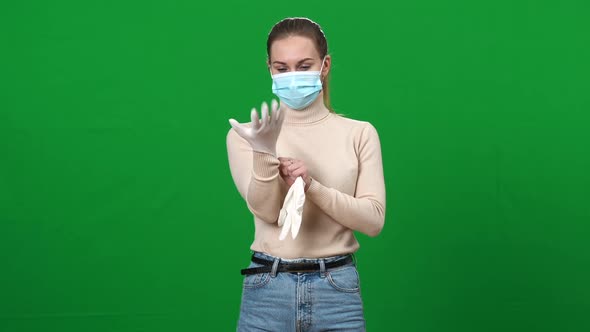 Concentrated Young Woman in Coronavirus Face Mask Putting on Protective Gloves Posing on Green