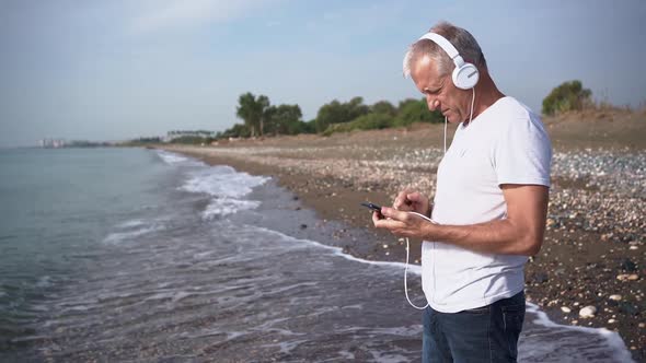 A Slender Whitehaired Elderly Man with Headphones Listens to Music on His Phone While Standing