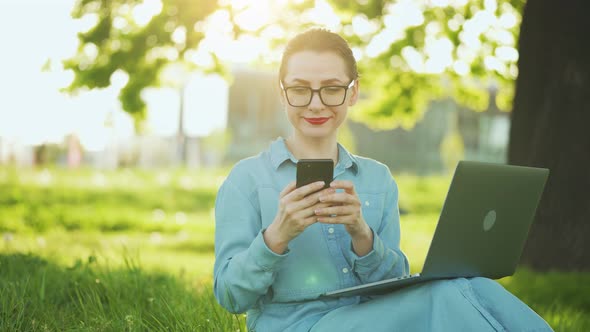 Busy Attractive Woman Working at the Laptop and Using Smartphone While Sitting on Grass in City Park